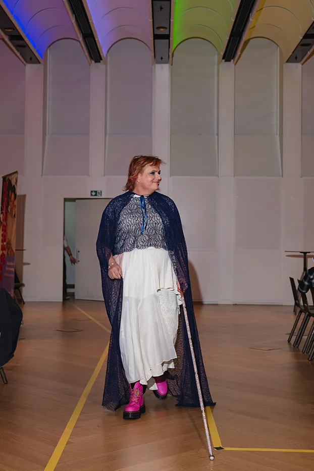 Model in weißem Kleid mit blauem Spitzenumhang und Blindenstock auf dem Laufsteg