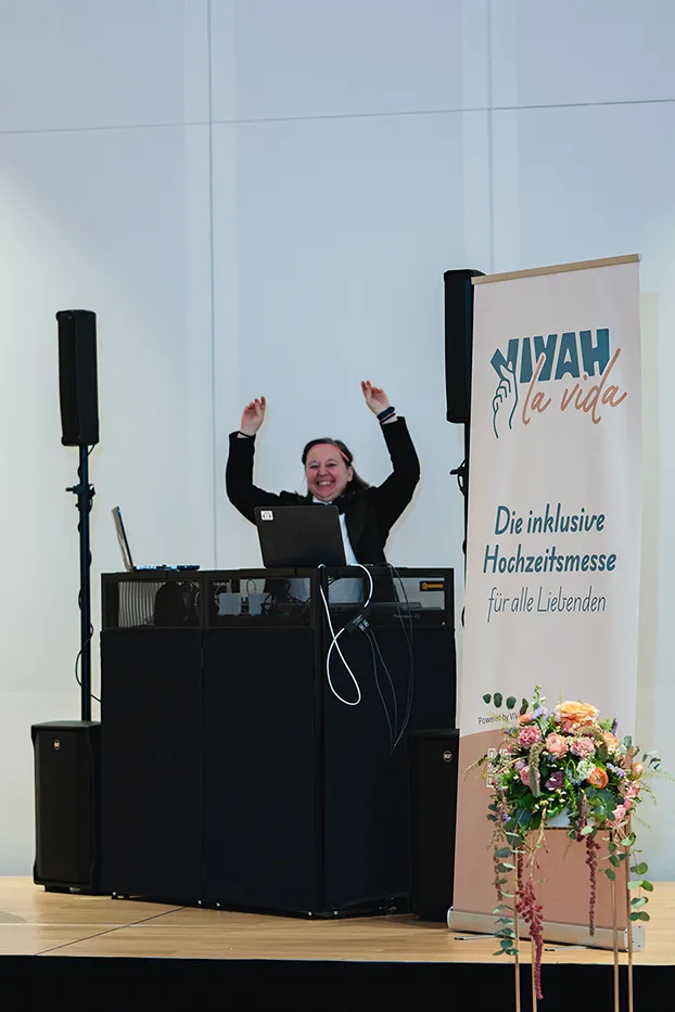 DJ hebt jubelnd die Arme hinter dem Pult neben dem Vivah la Vida Rollup