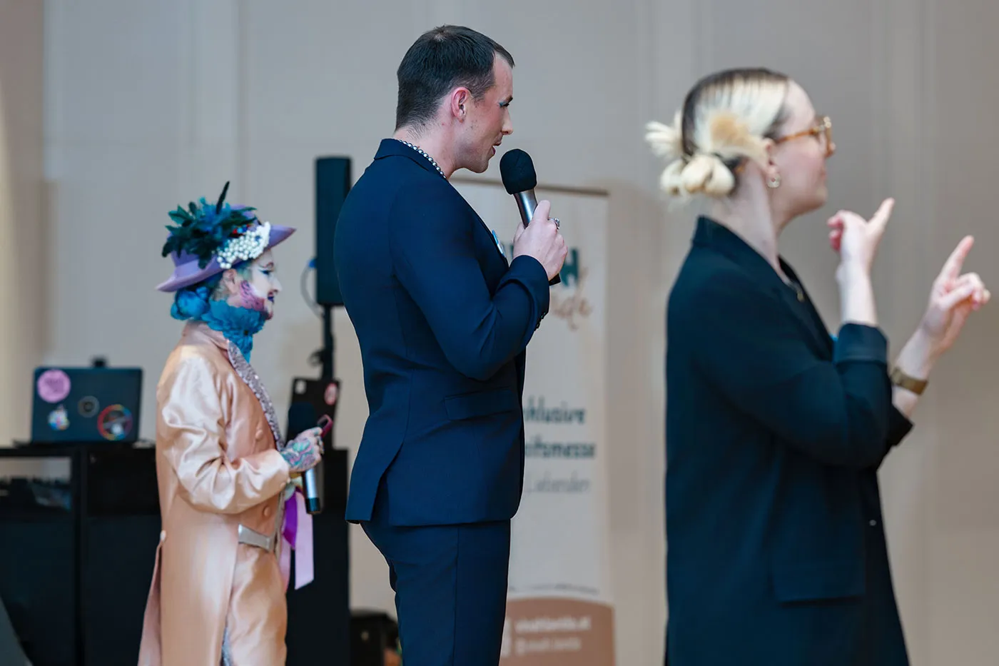 Moderator mit Mikrofon auf der Bühne, daneben eine Gebärdensprachdolmetscherin